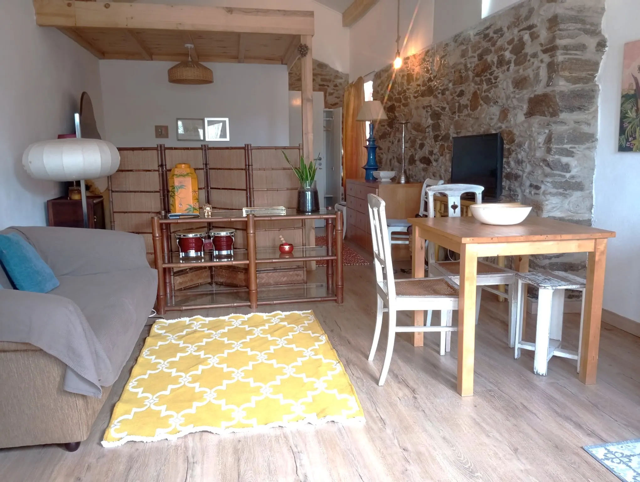 Open space da Casa Pequena: zona de estar, mesa de refeições e parede de pedra, sob o teto de madeira