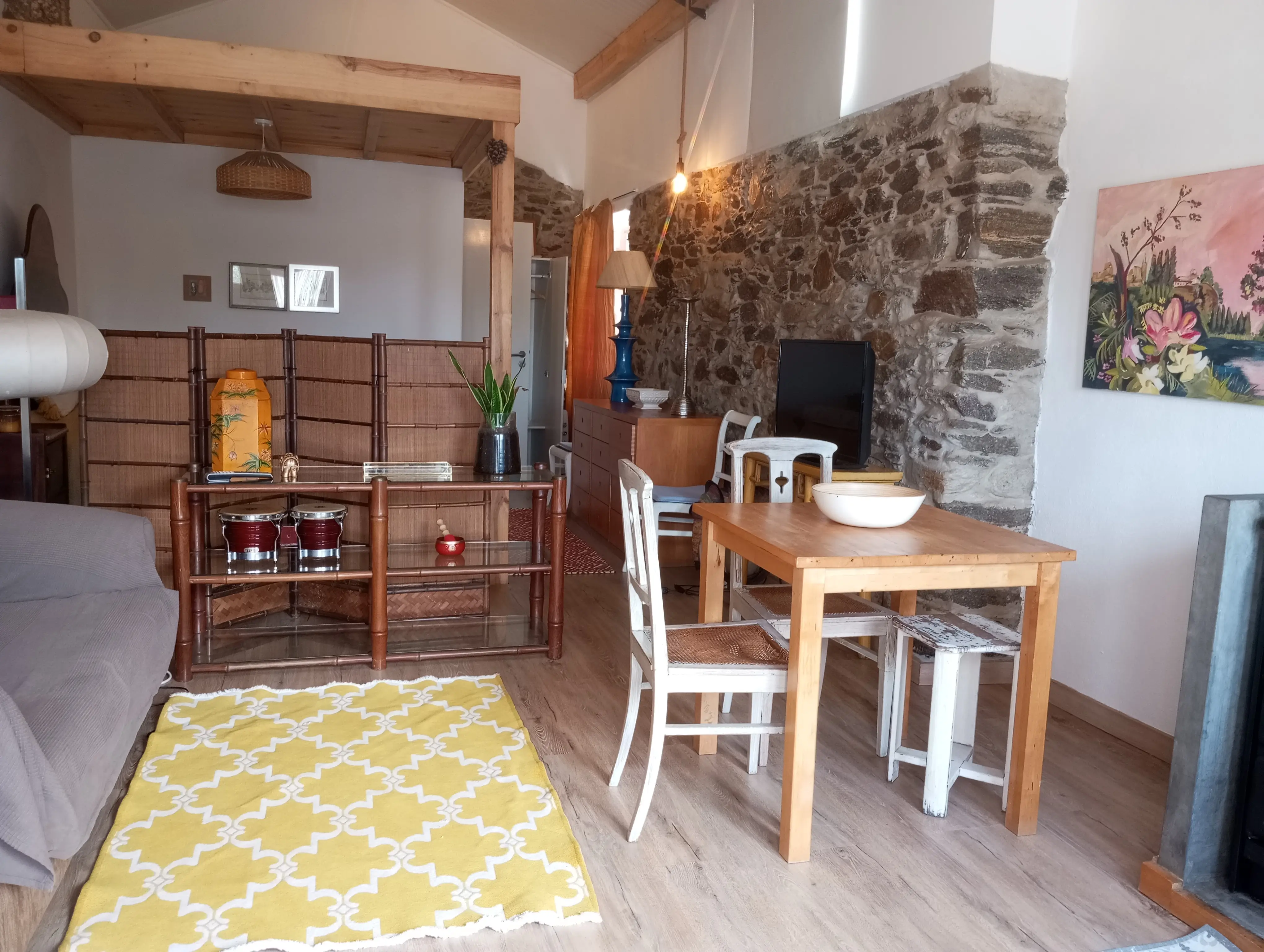 Sala da Casa Pequena com parede de pedra à vista, mesa de madeira, tapete amarelo e teto alto com vigas