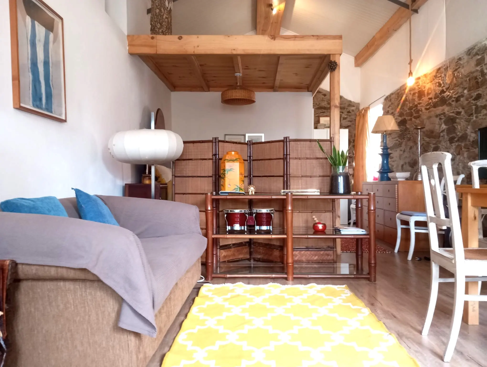 Sala com teto de vigas de madeira, parede de pedra e tapete amarelo, luz natural quente