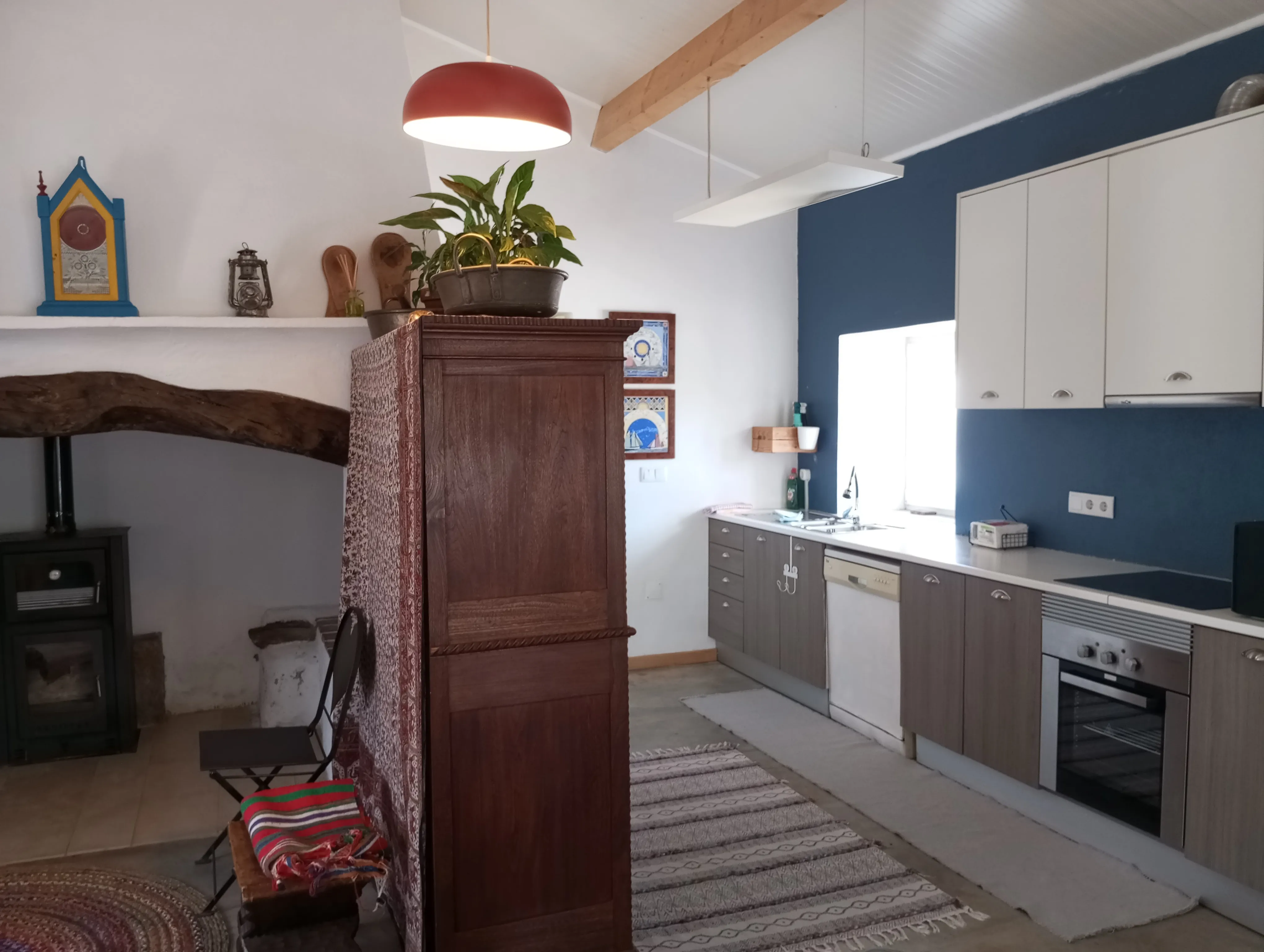 Sala e cozinha da Casa Principal em espaço contínuo, com a lareira antiga, o armário de madeira e a cozinha equipada de parede azul