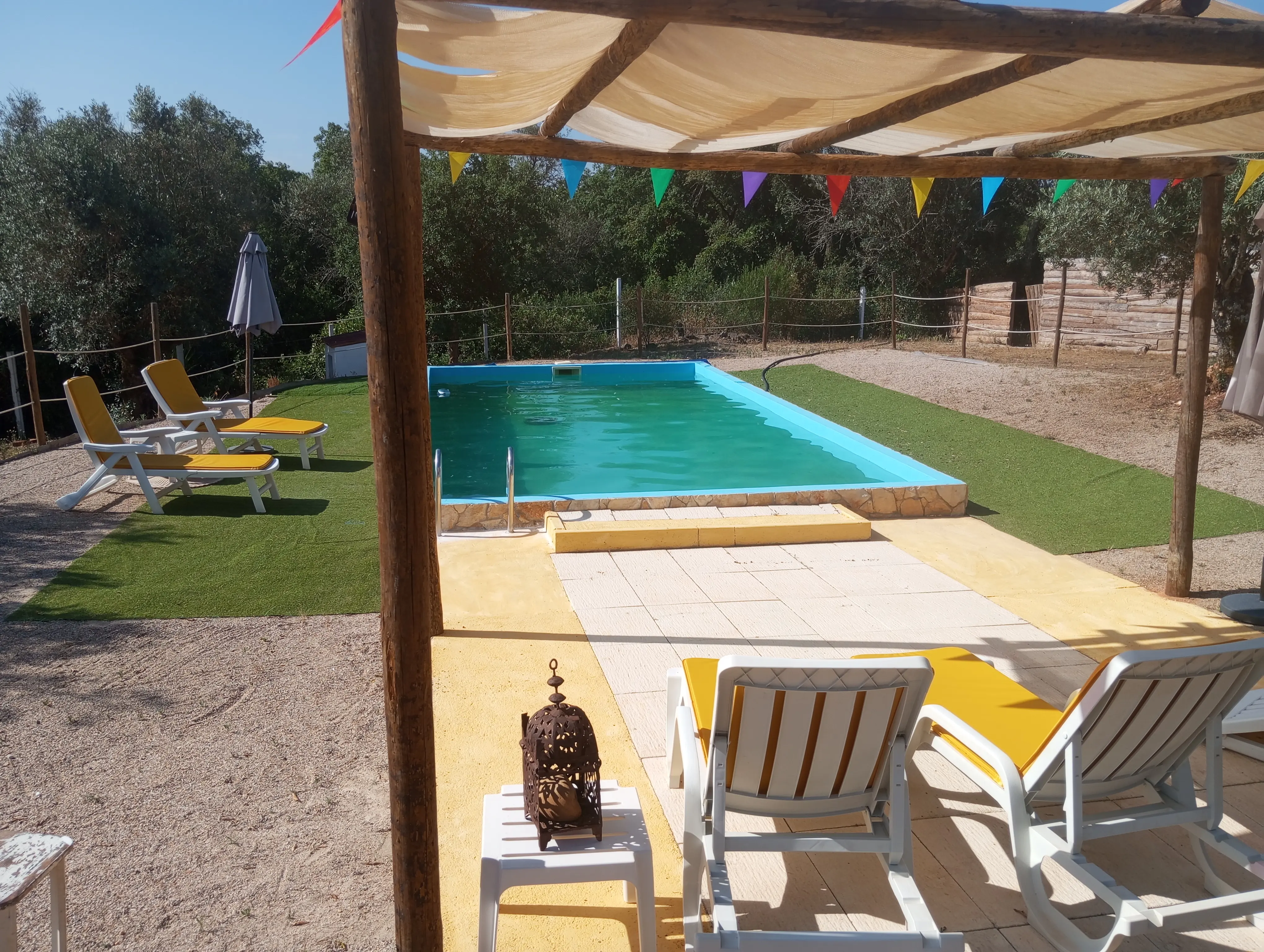 Piscina vista de baixo da pérgula de madeira, com espreguiçadeiras amarelas e bandeirinhas coloridas